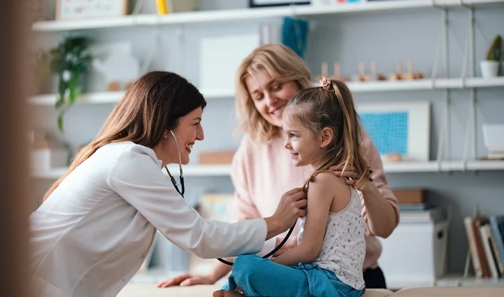 A friendly female pediatrician in a white coat examines a smiling young girl with a stethoscope during a medical check-up. The child's mother sits supportively behind her in a modern clinic office.