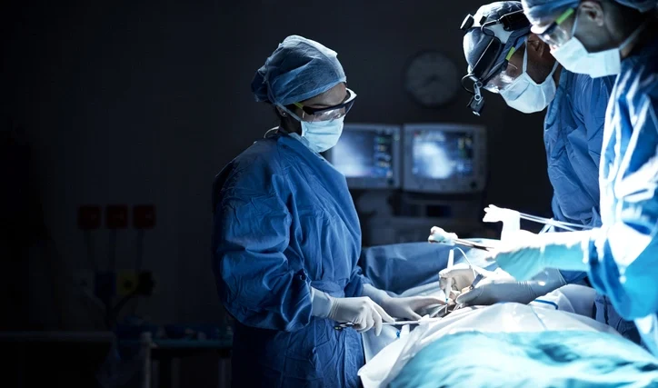 A friendly female pediatrician in a white coat examines a smiling young girl with a stethoscope during a medical check-up. The child's mother sits supportively behind her in a modern clinic office.