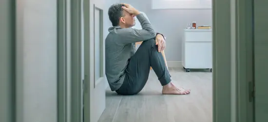A person sits barefoot on a wooden floor, seen through a doorway, holding their head in their hand. Their posture suggests stress, sadness, or a migraine. The image has a strong yellow-gold color filter.