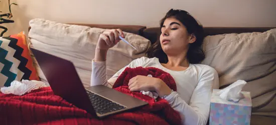 A young woman looking unwell lies on a couch under a blanket, checking a thermometer. A laptop, a box of tissues, and a hot water bottle are with her, suggesting she is sick with a cold or flu. The image is filtered with a heavy yellow-gold tint.