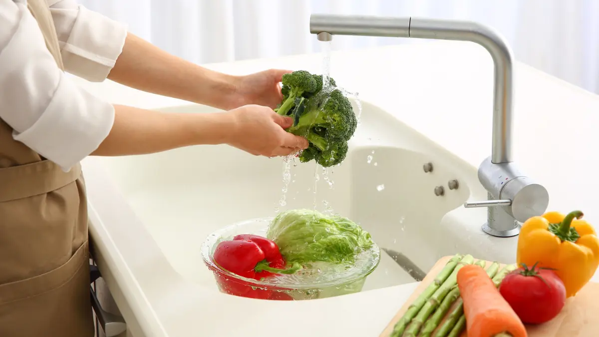 A person wearing a beige apron and white shirt washes a large broccoli floret with both hands under a stream of running water from a kitchen faucet, while a glass bowl of red pepper and lettuce and various other vegetables are nearby.