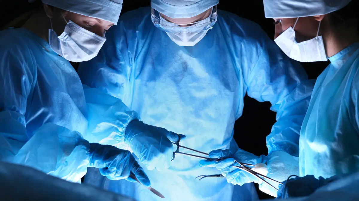 asc-vs-hospital-surgery-comparison Three surgeons in blue gowns, masks, and gloves perform an operation using surgical instruments.