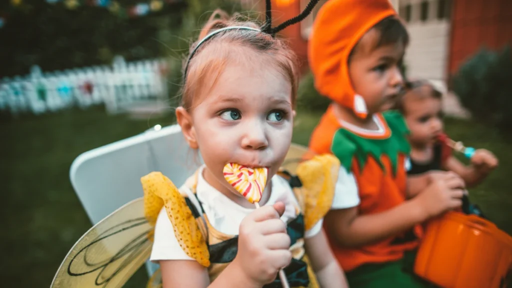 Young child in a bee costume eating a lollipop beside kids holding a pumpkin bucket of Halloween candy