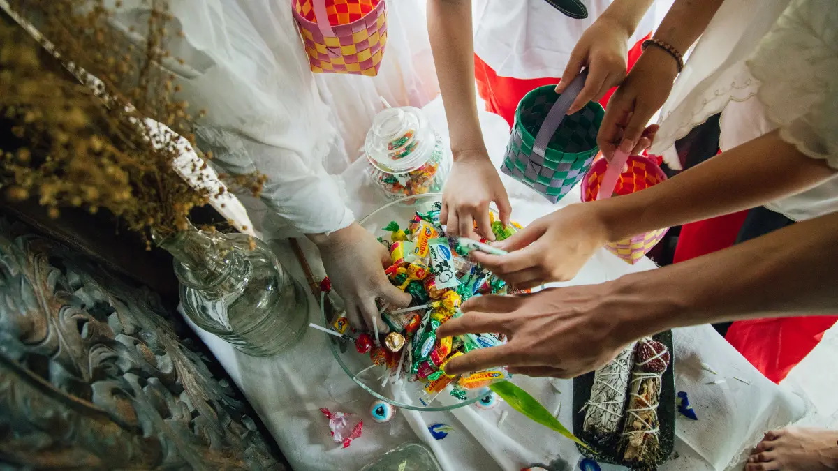 Kids' hands reaching into a glass bowl of wrapped leftover Halloween candy, holding woven treat baskets